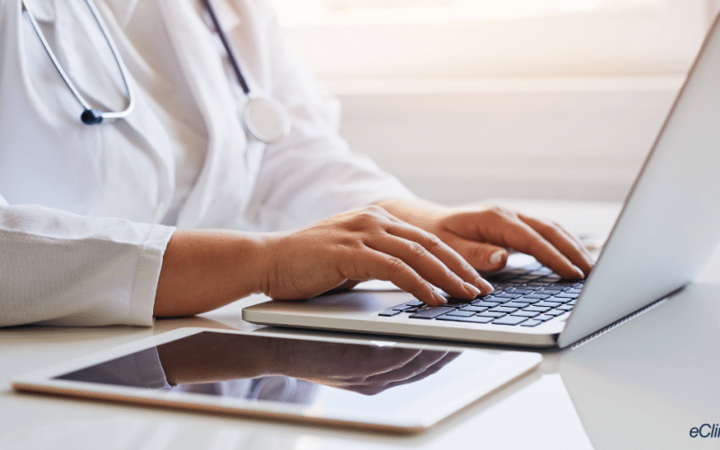 Doctor typing on laptop computer on desk with iPad next to the computer. eClinicalWorks logo in the lower right corner.